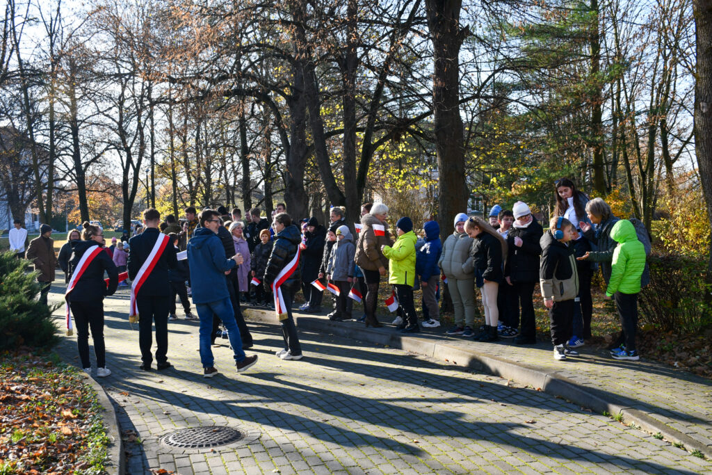 Grupa uczniów i nauczycieli stojących w parku podczas uroczystości patriotycznej.
