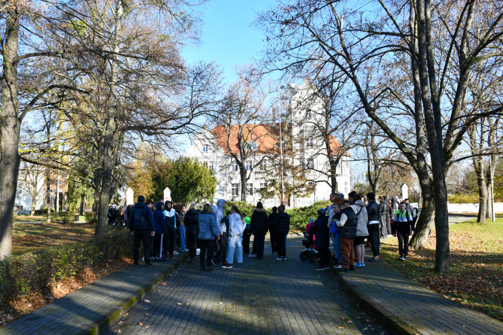 Grupa uczniów i nauczycieli stojących w parku podczas uroczystości patriotycznej. W tle budynek pałacu.