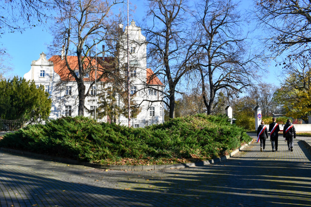 Młodzież z biało-czerwonymi szarfami trzymająca flagę podczas ceremonii. W tle budynek pałacu.