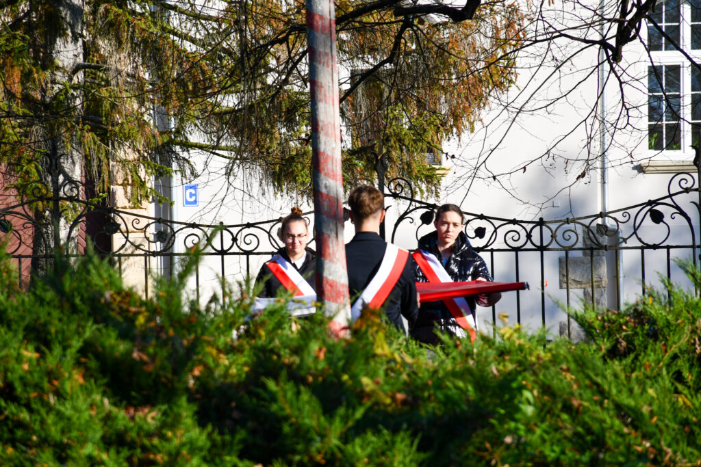 Młodzież z biało-czerwonymi szarfami trzymająca flagę podczas ceremonii.