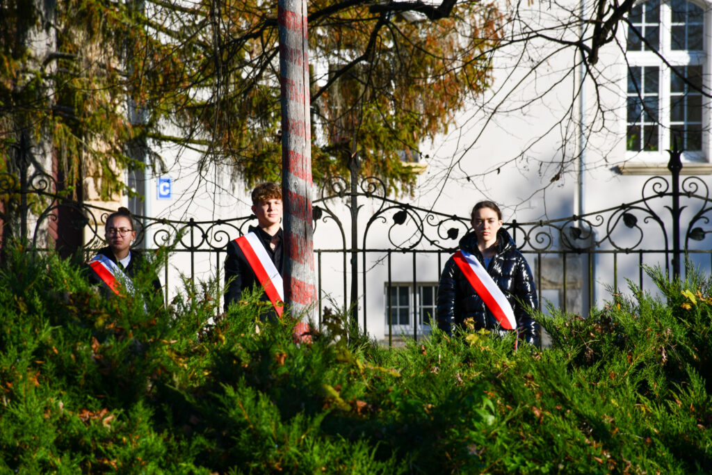 Młodzież z biało-czerwonymi szarfami stojąca za masztem na baczność.