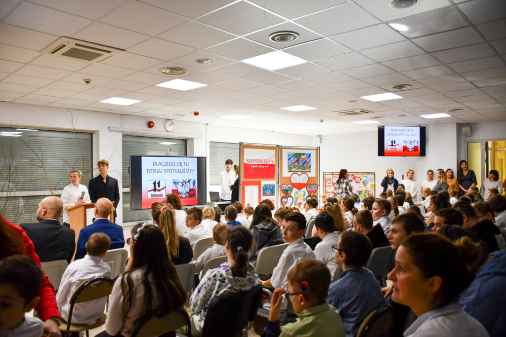 Publiczność siedząca na sali konferencyjnej podczas uroczystego apelu. W tle dekoracje patriotyczne.