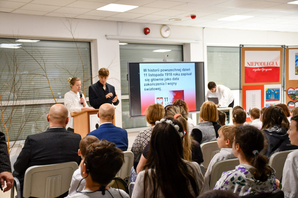 Uczniowie i nauczyciele siedzący na widowni podczas wystąpienia. W tle prowadzący i prezentacja.