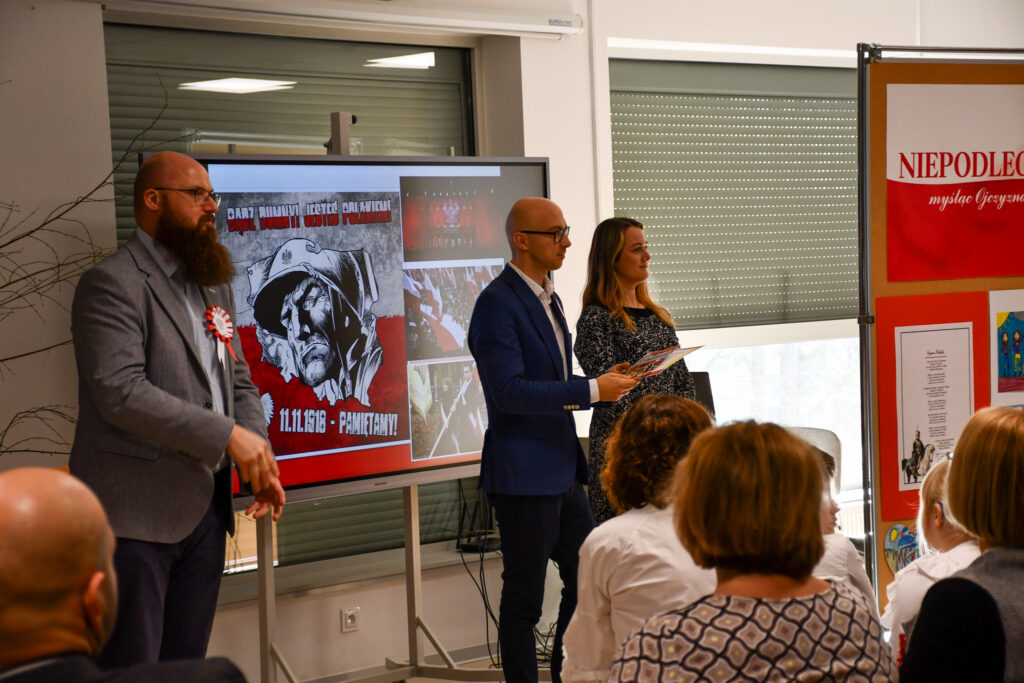 Prowadzący wydarzenie stojący przed ekranem z prezentacją historyczną.