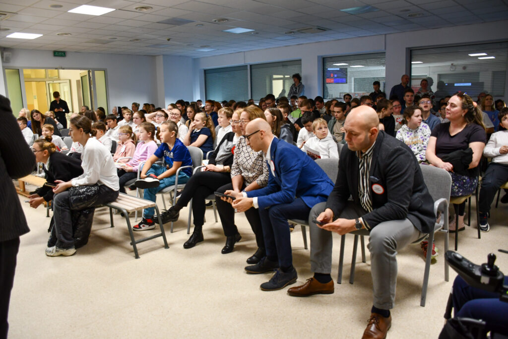 Widownia uczniów i nauczycieli siedzących podczas quizu multimedialnego z telefonami w dłoniach