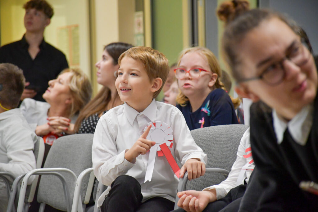 Uśmiechający się uczeń z przypinką w barwach narodowych siedzący wśród publiczności.