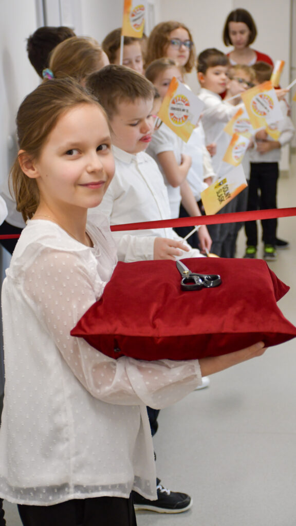 Dziewczynka trzyma czerwienną poduszkę z nożyczkami podczas uroczystego przecięcia wstęgi, w tle uczniowie z flagami.