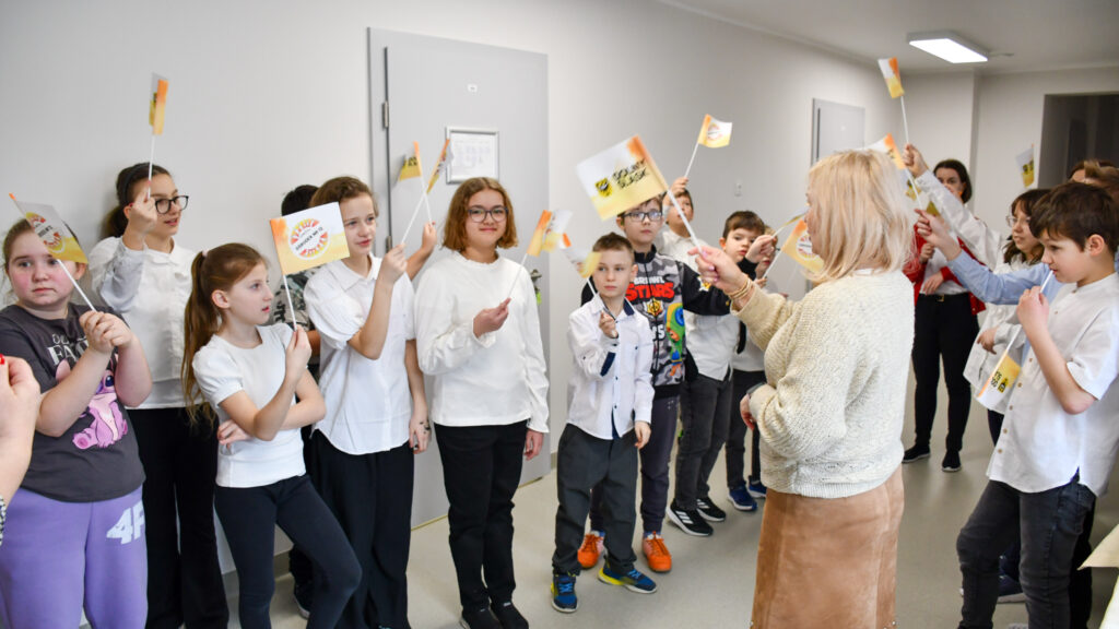 Grupa uczniów stoi w korytarzu szkolnym i macha małymi flagami podczas szkolnej uroczystości.