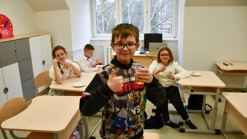 Uczeń pokazuje kciuk w górę, siedząc w klasie z napojem, w tle inni uczniowie przy ławkach.