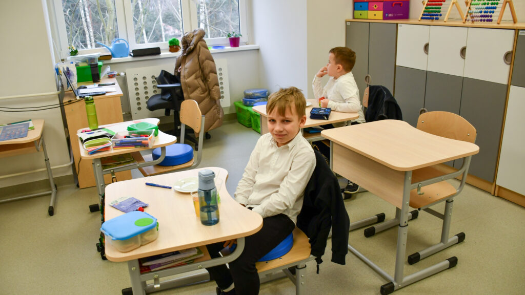 Uczeń siedzi przy ławce szkolnej z napojem, w tle inni uczniowie podczas przerwy.