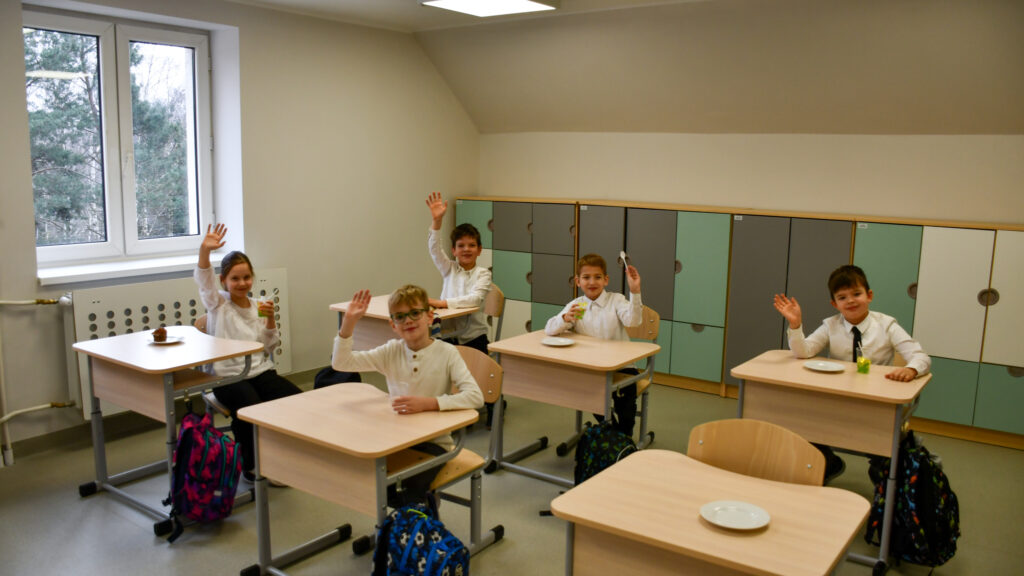 Uczniowie siedzą w klasie i podnoszą ręce, trzymając napoje podczas szkolnego spotkania.