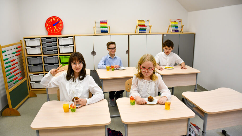 Grupa uczniów siedzi w klasie przy ławkach z poczęstunkiem i uśmiecha się do zdjęcia.