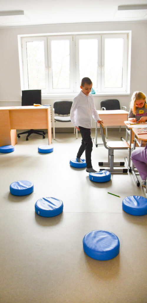 Uczeń przechodzi po sensorycznych poduszkach w sali lekcyjnej, w tle dzieci pracujące przy stoliku