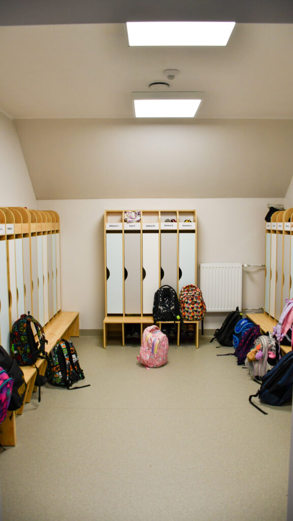 Szkolna szatnia z rzędami szafek i plecakami uczniów ustawionymi przy ławkach.