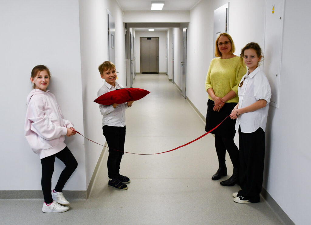 Dzieci i nauczycielka stoją w korytarzu z czerwoną wstęgą przed uroczystym otwarciem.