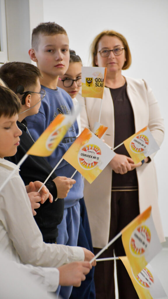 Uczniowie trzymają flagi podczas szkolnej uroczystości w obecności nauczycielki.