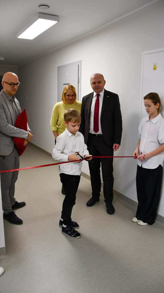 Uczeń przecina czerwoną wstęgę podczas szkolnej uroczystości w obecności nauczycieli i uczniów.