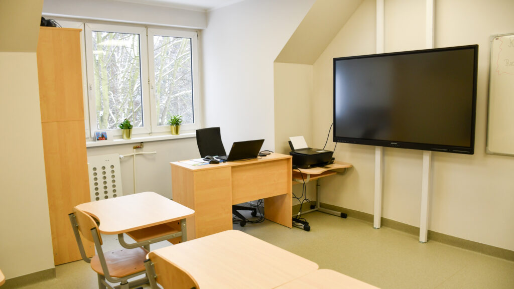 Stanowisko nauczyciela z komputerem i drukarką w wyposażonej klasie.