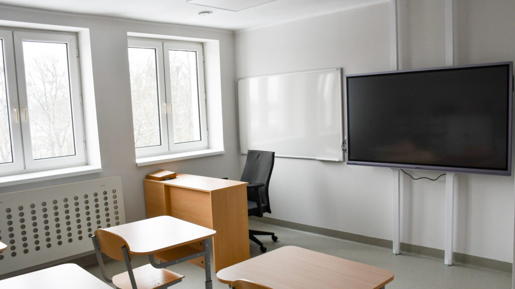 Sala lekcyjna z ławkami, biurkiem nauczyciela i tablicą multimedialną przy oknach.