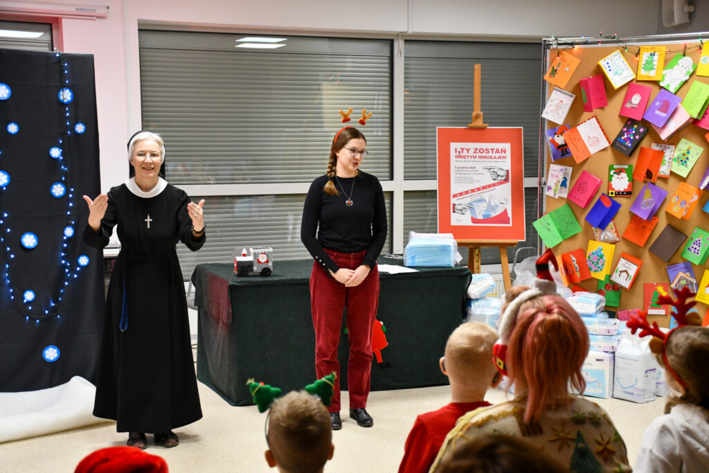 Siostra Zakonna i młoda kobieta stoją przed dziećmi podczas spotkania świątecznego, obok tablica z kartkami bożonarodzeniowymi.